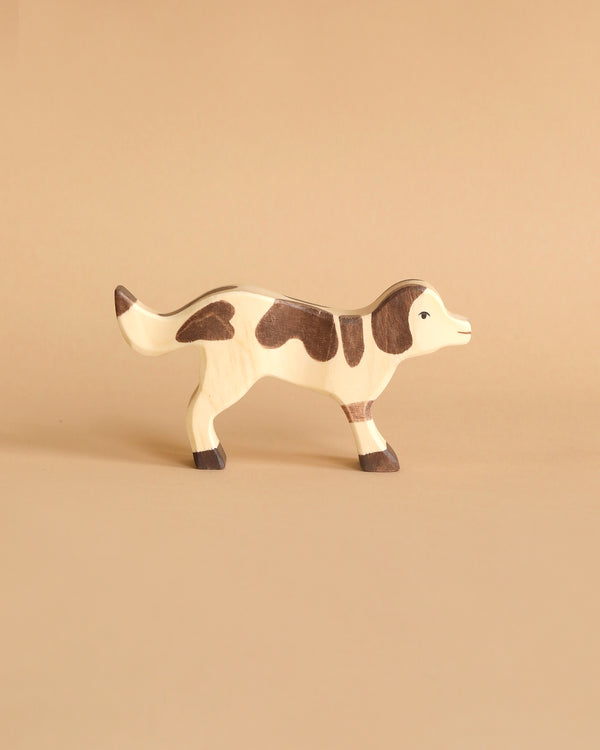 A small, wooden Holztiger Farm Dog handcrafted out of wood with a natural finish and dark brown markings stands on a light brown background. The simple design and smooth surface give it a minimalist and handcrafted appearance, characteristic of the high quality "Made in Europe" HOLZTIGER figures.