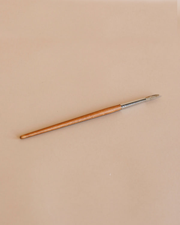 A single Cow Hair Paint Brush with a round tip, wooden handle, and metallic ferrule lies diagonally on a light beige background.