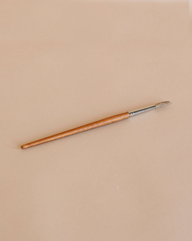 A single Cow Hair Paint Brush with a round tip, wooden handle, and metallic ferrule lies diagonally on a light beige background.