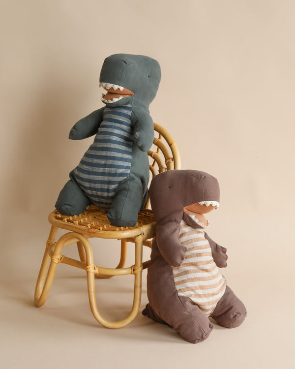 Two Maileg Large Dark Green Gantosaurus Dinosaurs sit against a beige background. One, in a blue and white striped shirt, stands on a small wicker chair. The other, in a brown and white striped shirt, sits on the floor beside the chair. Both toy dinosaurs have open mouths displaying their soft mix fabric teeth.