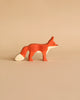 A small, handcrafted wooden **Holztiger Fox, Standing** with a reddish-brown body, white-tipped tail, and pointed ears stands on a plain beige background. This high-quality piece has a minimalist design, highlighting the fox's sleek, elegant silhouette reminiscent of HOLZTIGER figures.