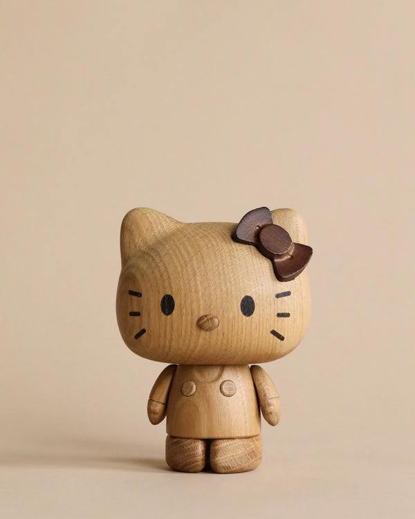 A small wooden figurine resembling Hello Kitty, Oak, crafted from European FSC White Oak. It features a large head, small body, and a bow on its right ear. The light brown figurine boasts minimalistic details with two black eyes, a nose, and whisker marks. Set against a plain beige background, it's an elegant addition to wooden contemporary design collectibles.