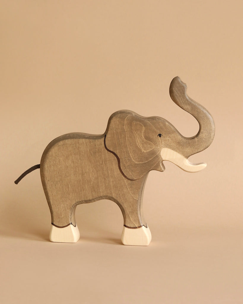 A Holztiger Elephant, Trunk Raised with a natural finish stands against a beige background. HOLZTIGER figures are known for their high-quality wood, and this handcrafted piece features white accents on its tusks, feet, and tail. The simple, minimalist design highlights the smooth texture of the wood.