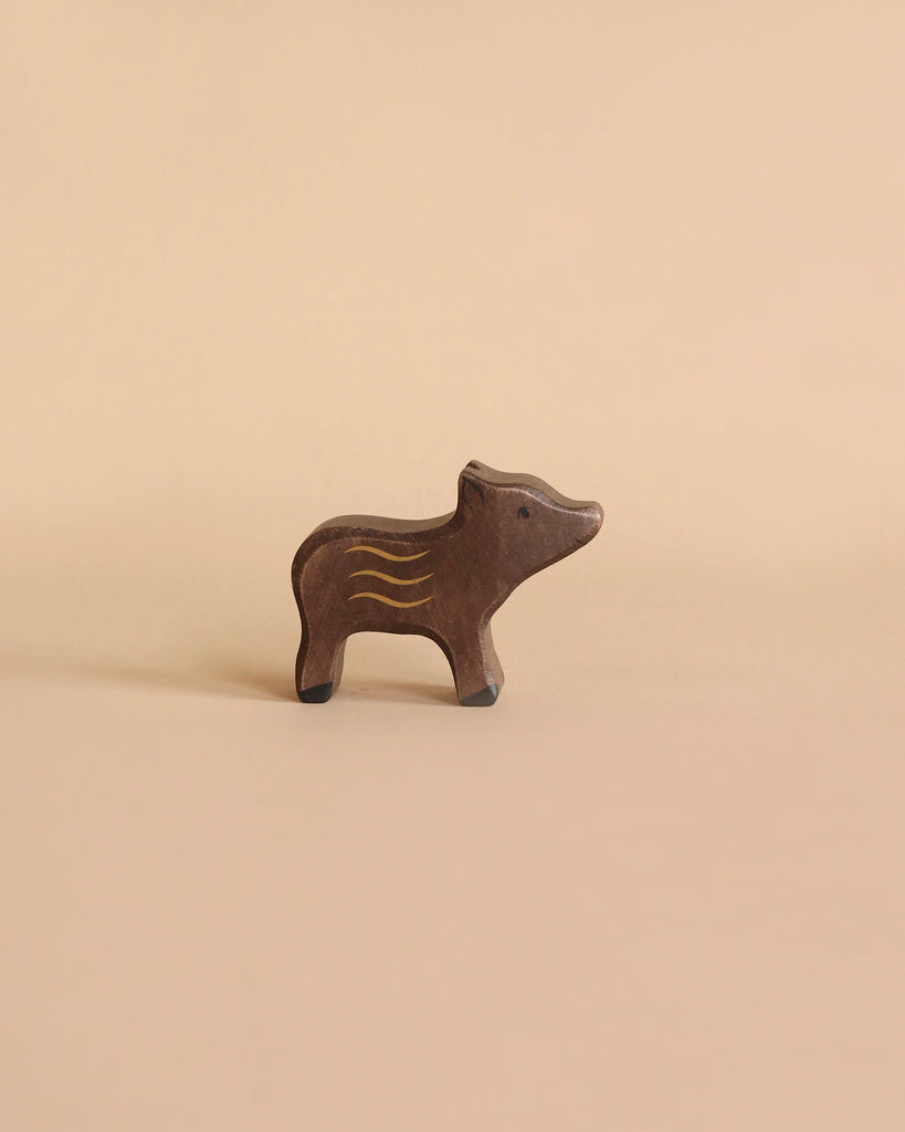 A small brown wooden figurine of a piglet with simple yellow and white markings on its body, set against a light brown background. This handcrafted wood piece by Holztiger Young Boar has a minimalist design, giving it a charming and whimsical appearance. Made in Europe.