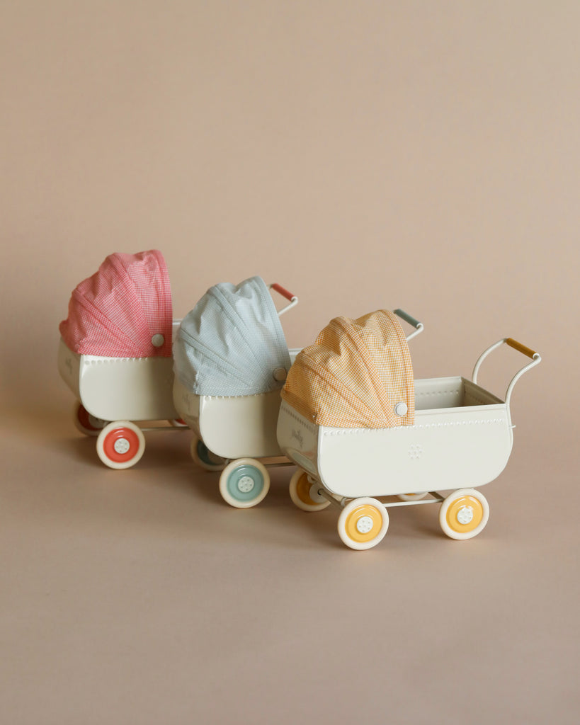 Three Maileg Micro Prams in a row, each with a unique colored fabric cover: pink, blue, and orange, against a soft beige background.