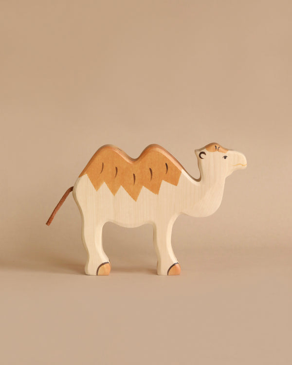 A simple, beige-colored wooden camel toy with dark brown accents on its back and orange highlights on its legs stands against a plain beige background. This charming piece is part of the Holztiger Camel collection, featuring a minimalist design with painted eyes and a slight smile, all handcrafted in Europe.