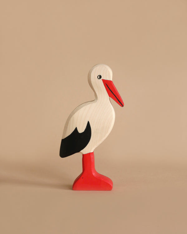 A handcrafted wooden Holztiger Stork stands against a plain beige background. The stork, part of the renowned HOLZTIGER figures, features a white body with a red beak, red legs, and black wingtips. Its simple design and smooth finish give it a whimsical and minimalist appearance. Made in Europe.