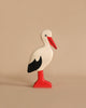 A handcrafted wooden Holztiger Stork stands against a plain beige background. The stork, part of the renowned HOLZTIGER figures, features a white body with a red beak, red legs, and black wingtips. Its simple design and smooth finish give it a whimsical and minimalist appearance. Made in Europe.