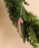 A delightful Maileg Christmas Metal Ornament - Sugar Cane - Stripe, hand-painted in red and white, hangs from a green pine branch, bringing a festive charm to the scene against a beige background.