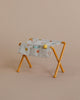 A Maileg Nursery Table, Baby Mouse - Ocher with mustard yellow metal legs is draped with a beige fabric featuring multicolored polka dots, perfect as a charming piece for a nursery table. The fabric is adorned with small round objects in yellow, green, and gray against a light brown background.