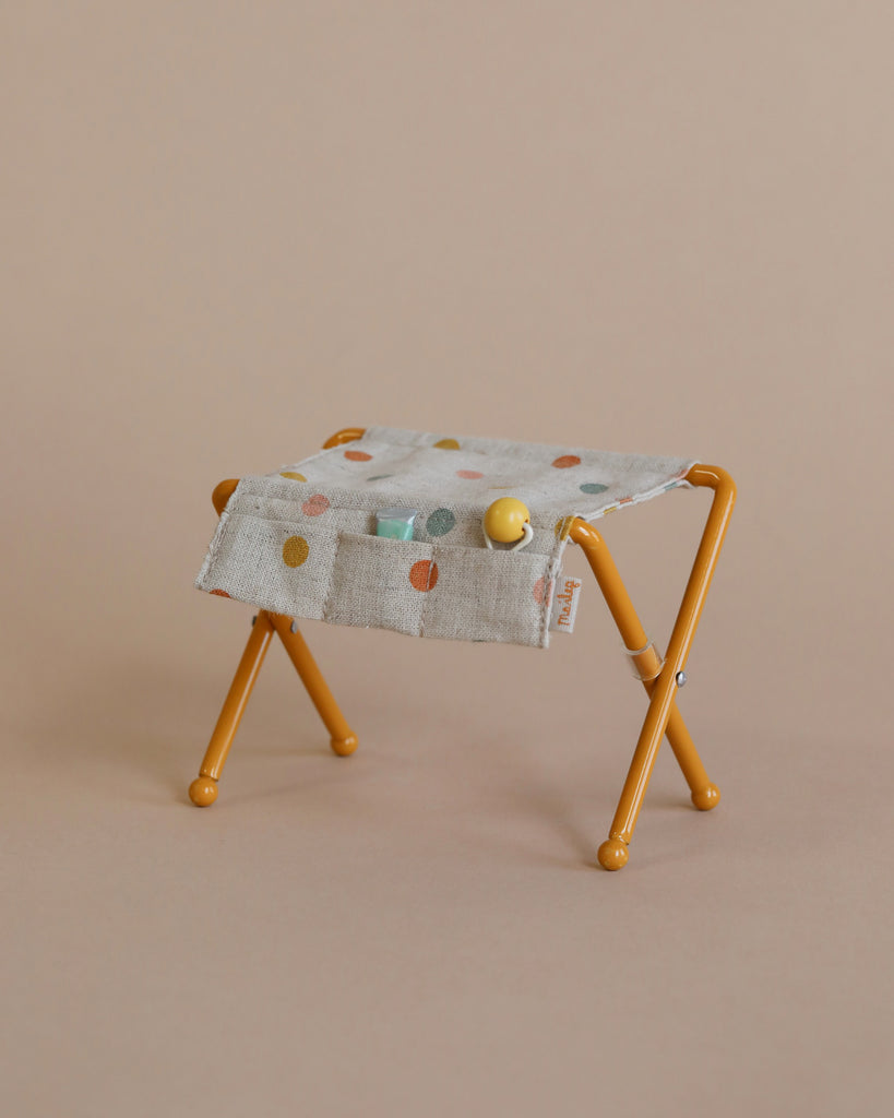 A Maileg Nursery Table, Baby Mouse - Ocher with mustard yellow metal legs is draped with a beige fabric featuring multicolored polka dots, perfect as a charming piece for a nursery table. The fabric is adorned with small round objects in yellow, green, and gray against a light brown background.