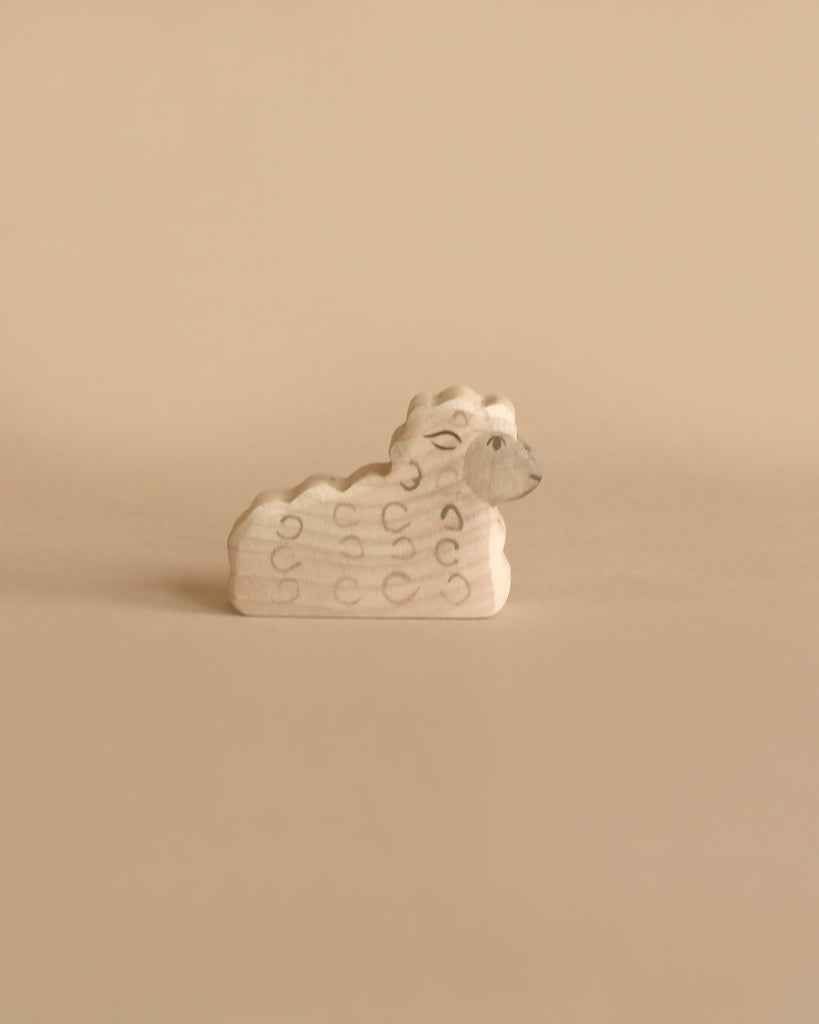 A small, handcrafted wood toy shaped like a sheep is set against a beige background. Made in Europe by HOLZTIGER figures, the Holztiger Lying Lamb features simple carved details including a curly wool pattern and a modest facial expression. The design is minimalistic, emphasizing natural wood tones.