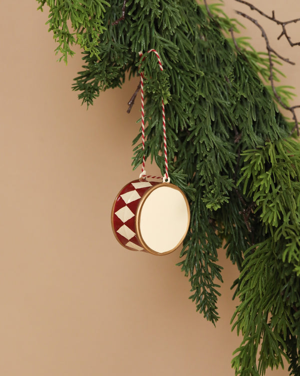 The Maileg Christmas Metal Ornament - Large Red Drum, featuring hand-painted red and white patterns, hangs from a matching string on a green fir branch against a beige background.