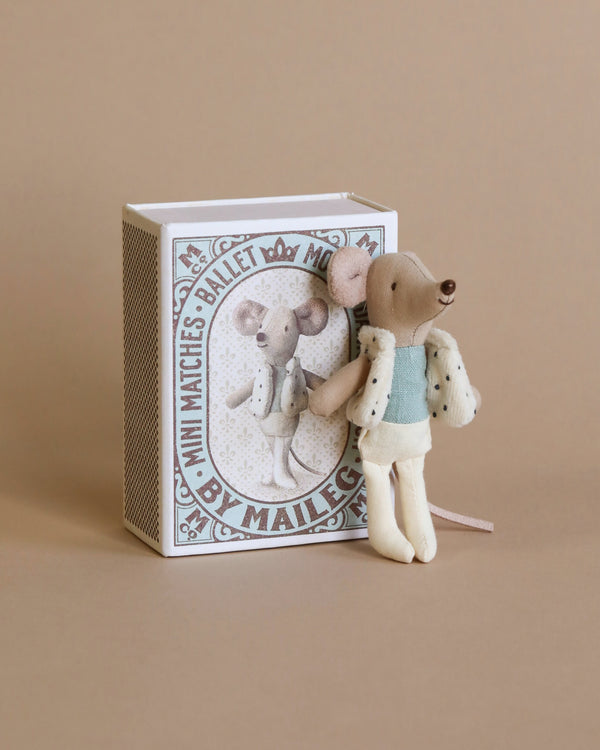 A small mouse doll dressed in a cute vest and light blue outfit stands next to a decorative matchbox featuring an illustration of a similarly dressed dance mouse. The box has the text "Maileg Little Brother Dancer Mouse in A Box." The background is a plain, light beige color.