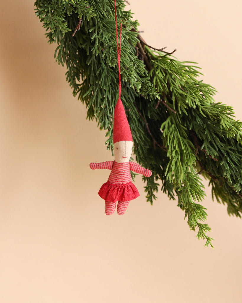 The Maileg Pixy Ornament - Girl is a small, red fabric doll with a pointed hat, hanging gracefully from a green pine branch. Its red striped outfit features a skirt and arms spread wide, creating an exclusive look against the beige background—perfect for adorning your Christmas tree.