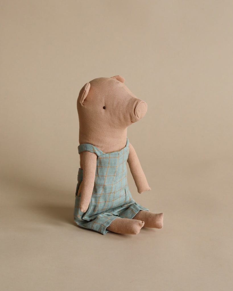 A handmade Maileg Pig In Overalls sits on a plain beige background. The adorable pig is dressed in a light blue plaid romper with yellow lines. The simple, minimalist design highlights the pig's rounded snout and floppy ears. This weighted stuffed animal is positioned in a seated posture.