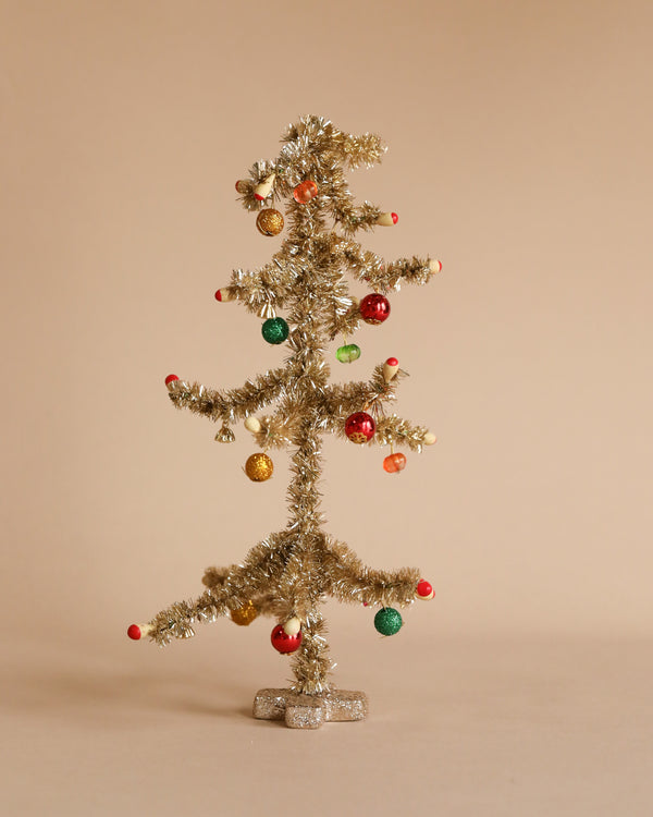 A small, sparse gold Christmas tree from the Maileg collection features vibrant red and green ornaments and stands elegantly against a plain beige background.