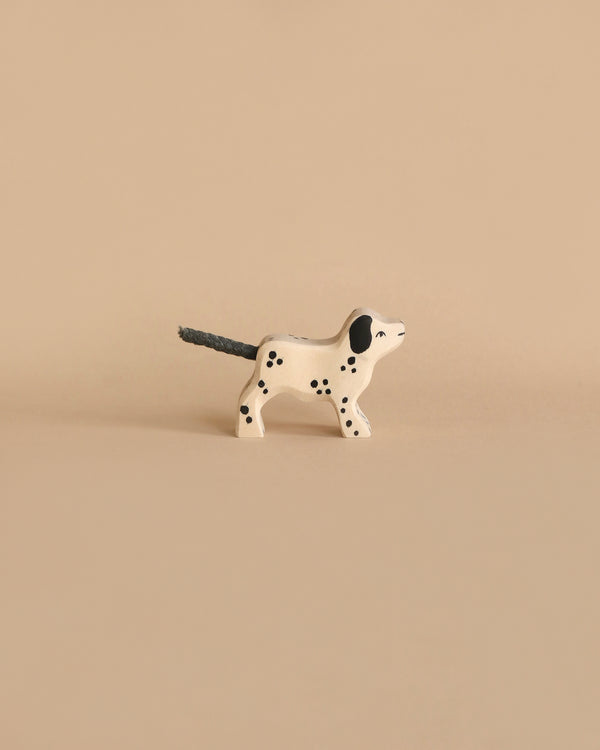 A small, minimalist wooden dog figurine painted white with black spots stands against a beige background. Handcrafted in Europe, this Holztiger Dalmatian Dog, Small figure's tail is black and slightly elevated. The scene is simple and uncluttered, focusing solely on the artisanal dog figurine.