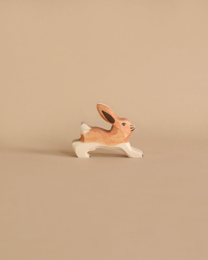 A handcrafted wooden toy rabbit painted in natural brown and white colors is shown against a beige background. The rabbit is depicted in a running pose with its ears upright and a small tail. Crafted from maple and beech wood, the delicate details of this Holztiger Rabbit, Running figure create a charming and whimsical appearance.