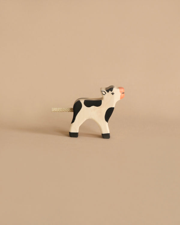 A small wooden toy cow with black and white patches stands on a plain beige background. Handcrafted in Europe, this Holztiger Black Calf figure features simple, minimalist details, including a short, rope-like tail and an orange snout.