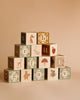 Introducing the Uncle Goose Watercolor ABC Blocks: a set of artistically designed blocks crafted from sustainable basswood, featuring letters and delightful illustrations such as animals, a mushroom, and a strawberry on a beige background. These blocks display decorative patterns in soft colors, using safe-to-touch inks. Perfect for building creative pyramids!