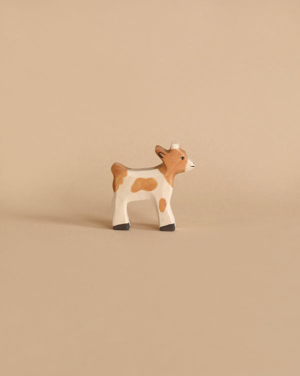 A small, handcrafted wooden figurine of a calf, painted white with light brown patches, black hooves, and a short tail, stands on a solid beige background. Made in Europe by Holztiger Baby Goat.