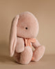 The Maileg Medium Plush Bunny - Powder, a soft and plush toy with long ears, crafted from gentle fabric and polyester fillings, is adorned with a ribbon around its neck as it sits against a plain beige background.