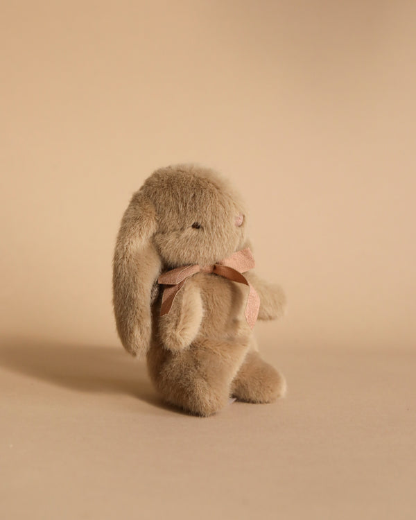 The Maileg Mini Plush Bunny in Dusty Brown, crafted from soft fabric and filled with polyester for ultimate cuddliness, features floppy ears and a pink bow around its neck as it sits upright on a plain beige background.