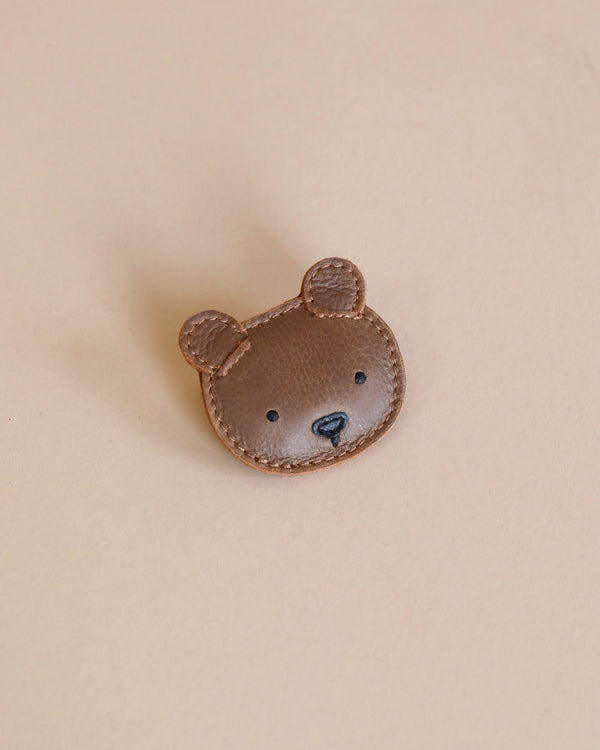 A cute Donsje Leather Hair Tie - Bear face patch with stitched details on a beige background. The bear has small, round ears and a black nose and mouth.