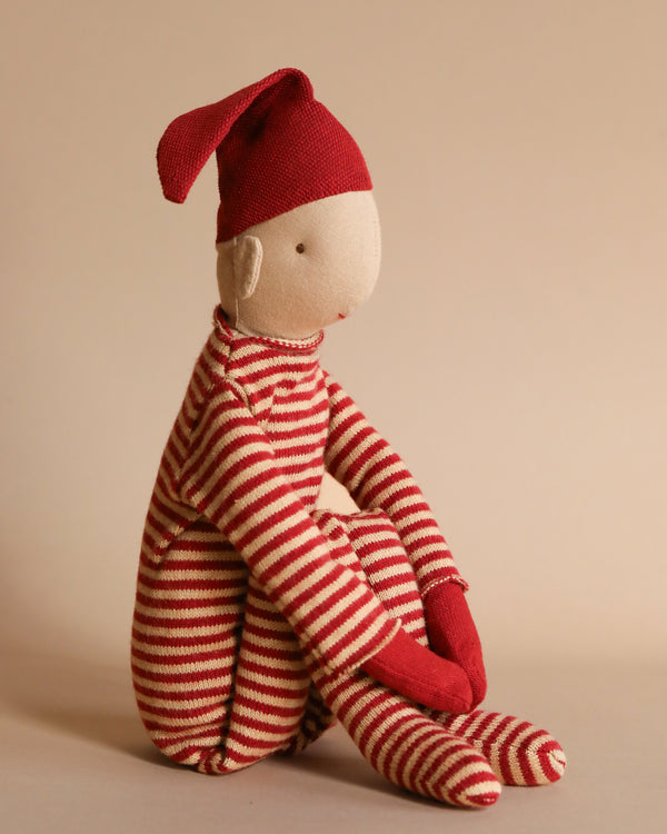 The Maileg Long Legged Pixy, Large is a plush fabric doll in a sitting position wearing a red and white striped outfit, complete with a matching pointed hat and red mittens, capturing the whimsical charm of a pixy. The doll is set against a plain, neutral background.