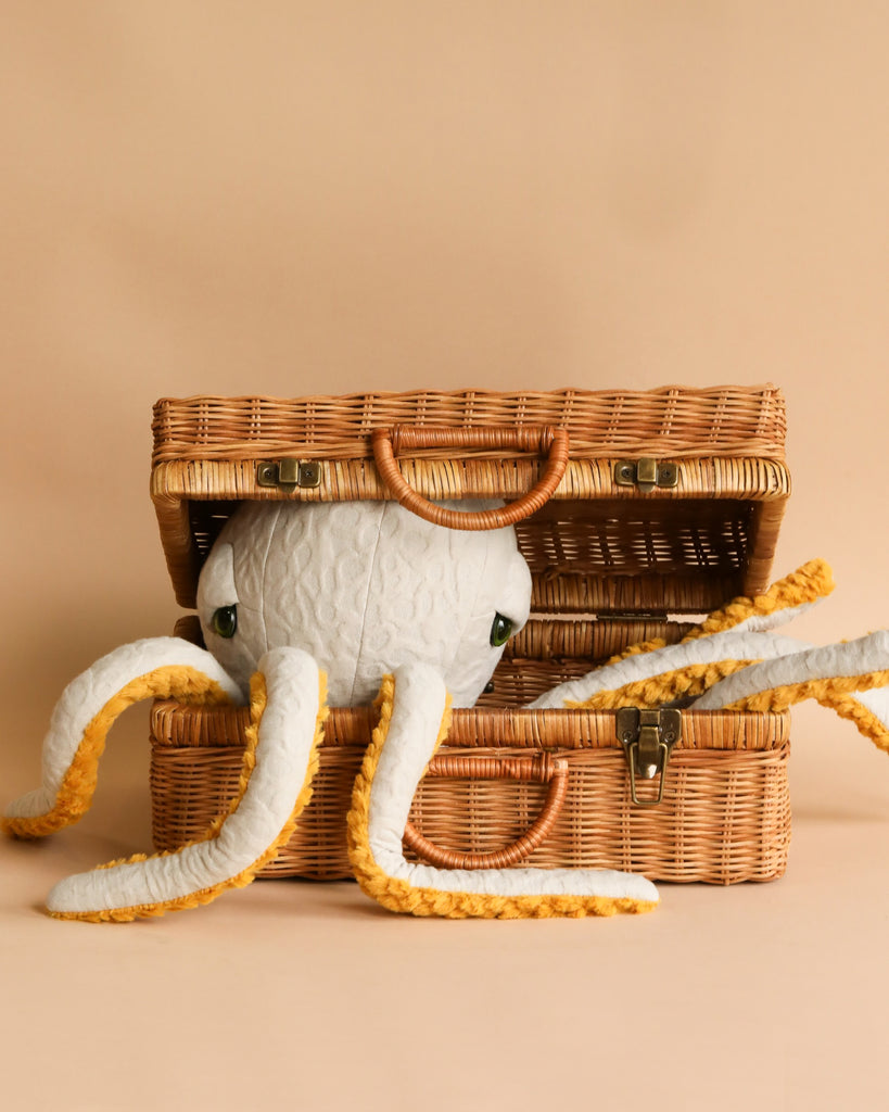 A BigStuffed Octopus - Pop with fluffy white tentacles edged in yellow rests inside an open, vintage-style wicker suitcase. The background is a warm beige color, complementing the suitcase and creating a soft, inviting scene reminiscent of an underwater world.