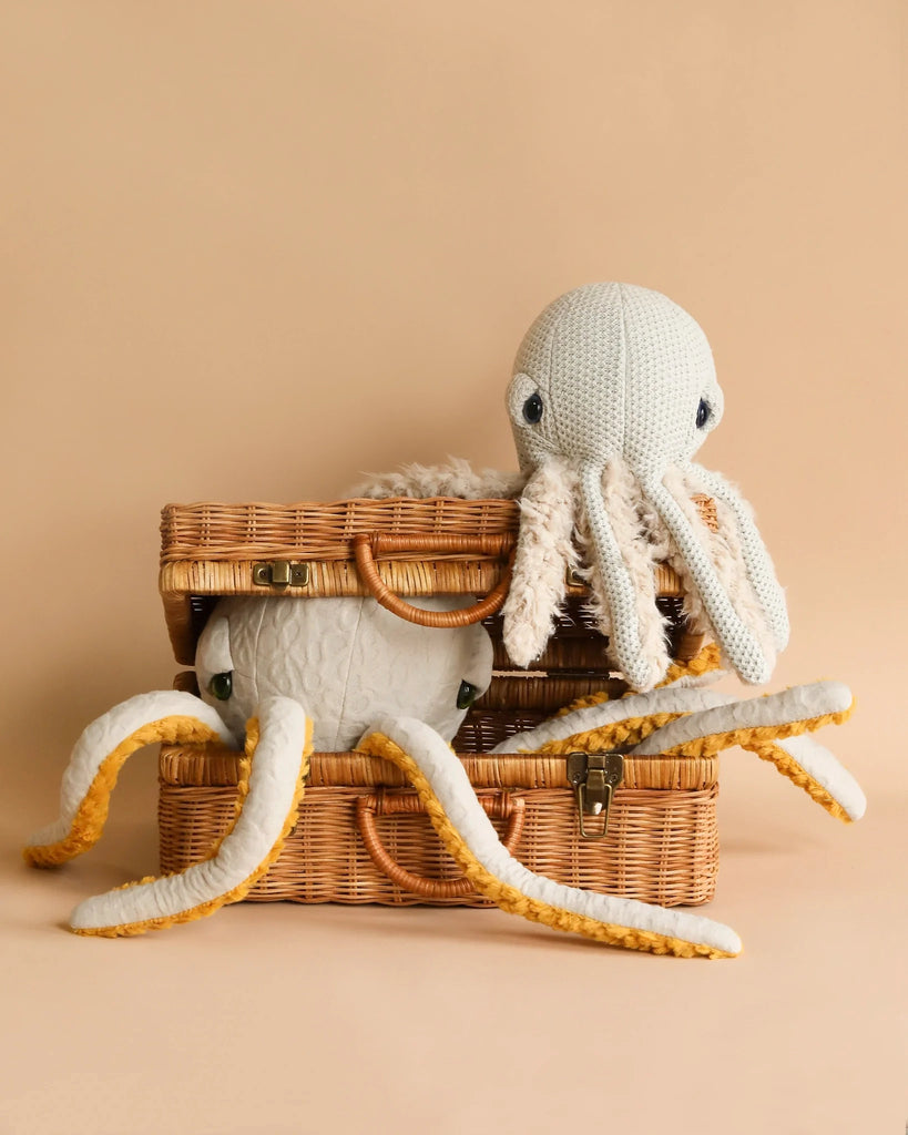 A BigStuffed Mini Grandma Octopus with a white and yellow color scheme is seen sitting on top of a wicker basket, while another mini plush octopus peeks out from an open wicker basket below it. The background is a plain, soft peach color.