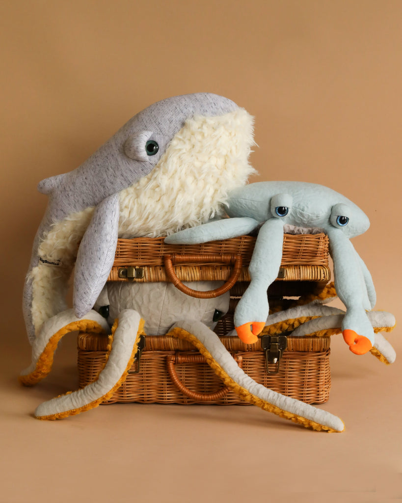 Two large stuffed animals, a BigStuffed Small Grandpa Whale and an octopus, rest on top of two stacked wicker baskets against a beige background. The whale has a soft, textured body with a white underbelly, and the octopus has light blue arms with orange tips. Ideal children's companions for cozy moments.