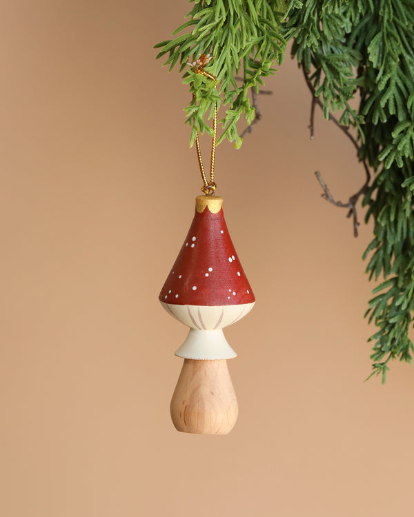 A Handmade Wooden Mushroom Ornament, standing 4 inches tall with a red and white cap, hangs from a pine branch against a solid beige background.