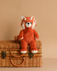 Benji the red panda, a plush toy with a red and white fur pattern and a friendly expression, sits on a closed wicker basket against a beige background. This adorable Steiff, Benji Red Panda Stuffed Animal Plush, 11 Inches even features their signature Button in Ear, making it perfect for collectors and kids alike.