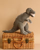 A Steiff, Thaisen Tyrannosaurus "T-Rex" Dinosaur Plush Toy, 18 Inches with felt teeth and spikes stands on top of a closed wicker basket with leather handles and brass clasps, set against a neutral beige background.