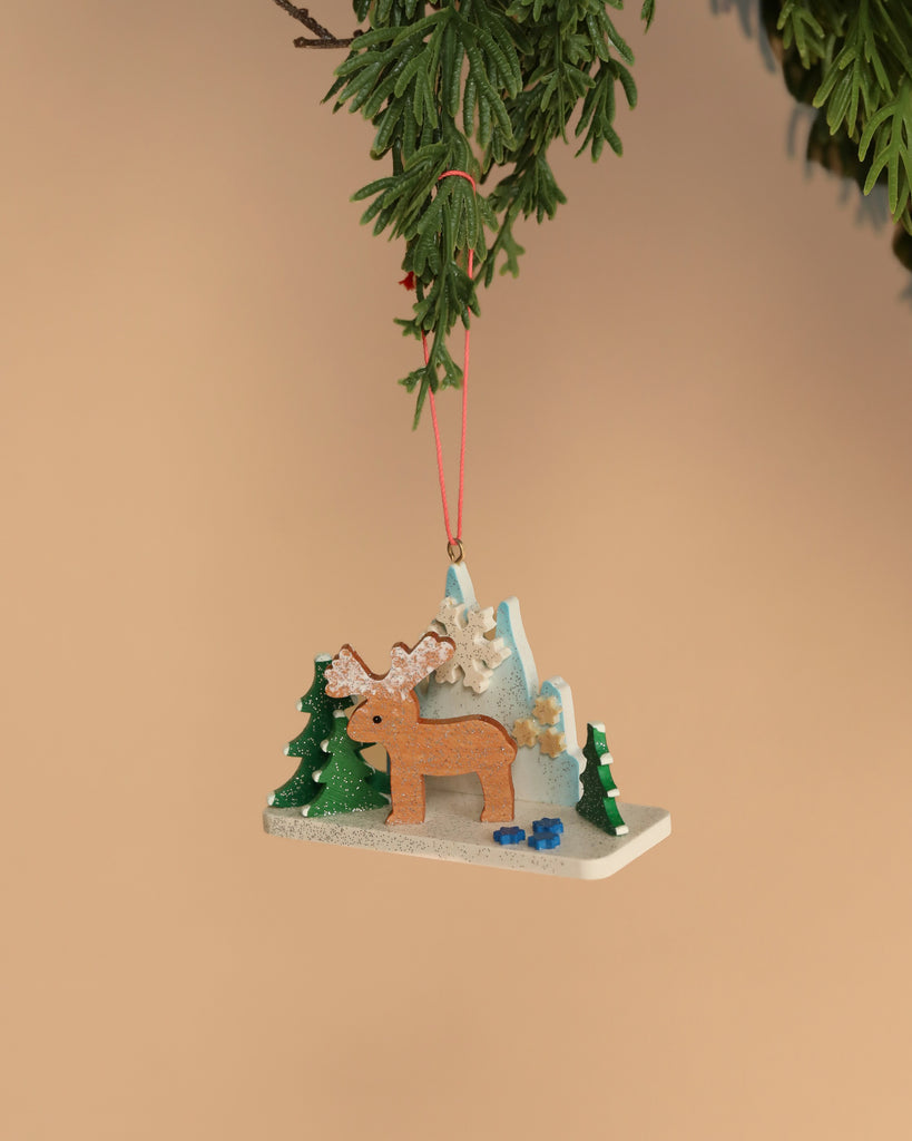 A festive Christian Ulbricht Ornament, crafted in Germany, hangs from a branch, showcasing a reindeer with antlers amidst snow-covered trees. In the background, a snowflake is set against a cream-colored backdrop.