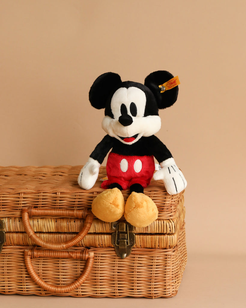 A Steiff, Disney's Mickey Mouse Stuffed Plush Toy, 12 Inches, showcasing Steiff craftsmanship with its big black ears, white gloves, red shorts, and yellow shoes, sits atop a closed woven wicker basket with leather straps and metal clasps against a beige background.