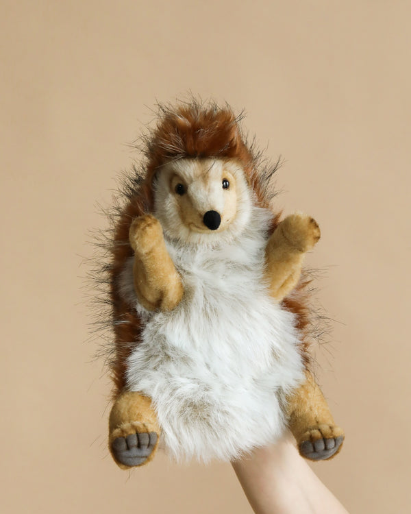 A hand is holding the Hedgehog Puppet, showcasing its realistic features against a beige background. The adorable creature, reminiscent of HANSA animals, displays brown and white fur, tiny black eyes, and a small black nose.