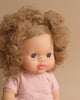 A Minikane Baby Doll (13") - Anais with curly light brown hair and big blue eyes wears a pale pink dress, exuding a gentle natural vanilla scent. The background is a plain beige color.