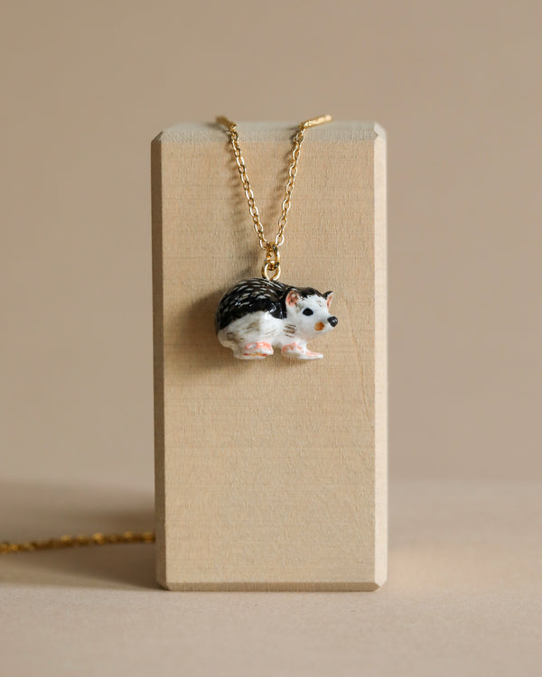 A Hedgehog Necklace with a hand painted pendant shaped like a small hedgehog resting on a wooden display block. The pendant’s black and white detailing gives it a realistic appearance, complemented by the simple, neutral beige background.