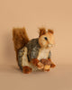 The Sitting Gray Squirrel Stuffed Animal, featuring a bushy tail and realistic characteristics, is positioned against a beige backdrop, clutching an acorn in its front paws. This handcrafted plush toy showcases intricately sewn fur patterns in brown, gray, and white hues.