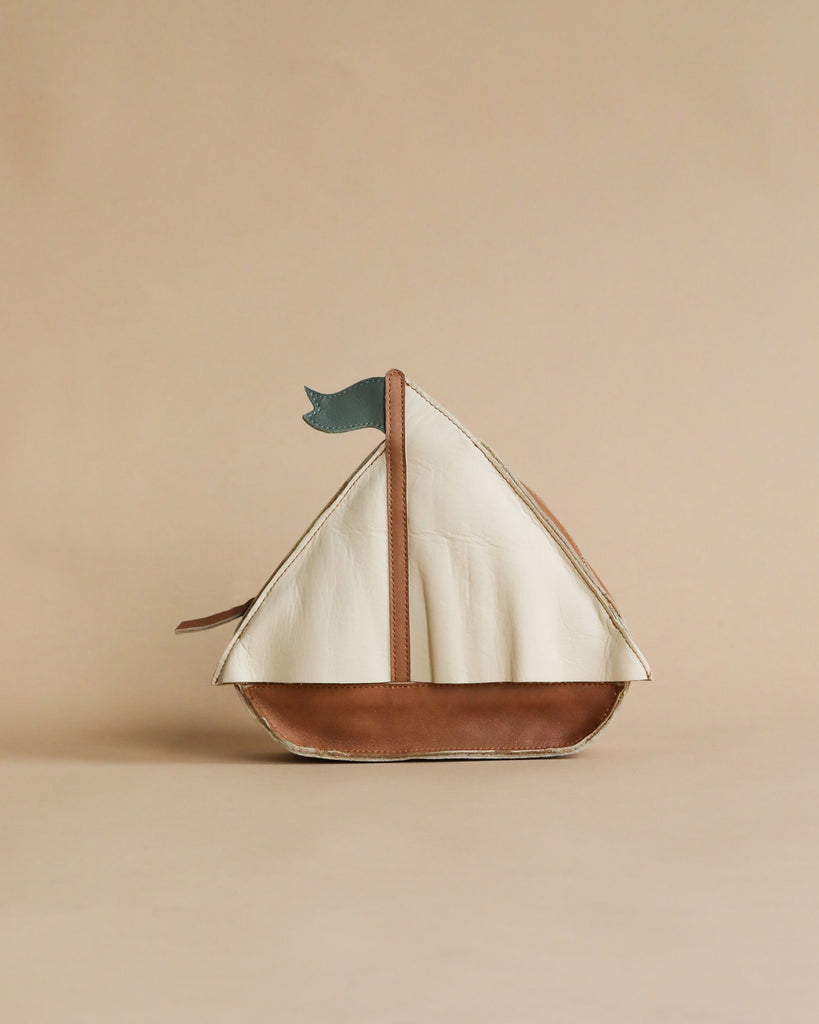 A Donsje Mini Leather Backpack - Boat with a premium leather base and white sails, adorned with a small blue flag, standing against a plain beige background.