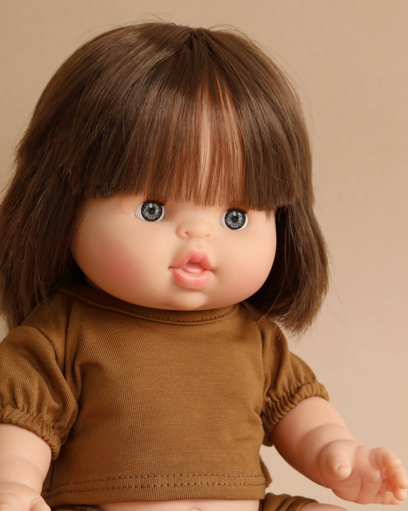 A Minikane Baby Doll (13") - Chloe with straight brown hair and bangs, dressed in a brown outfit. The doll has blue eyes, rosy cheeks, and pink lips with a natural vanilla scent. The background is a plain beige color.