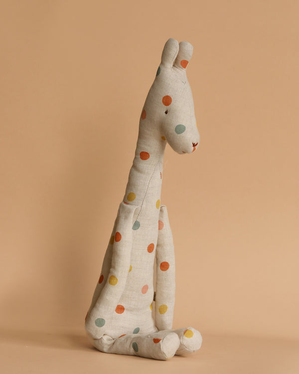 The Maileg Medium Giraffe is an upright plush toy made from recycled polyester, featuring a light beige fabric with vibrant polka dots in red, blue, yellow, and green. This safari companion rests beautifully against a warm beige backdrop.