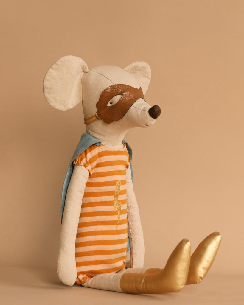 The Maileg Superhero Mouse, Maxi - Girl is dressed as a superhero in an orange striped outfit and a blue cape, paired with a brown mask. Against a beige backdrop, it features large ears and shiny gold shoes, all made from recycled polyester.