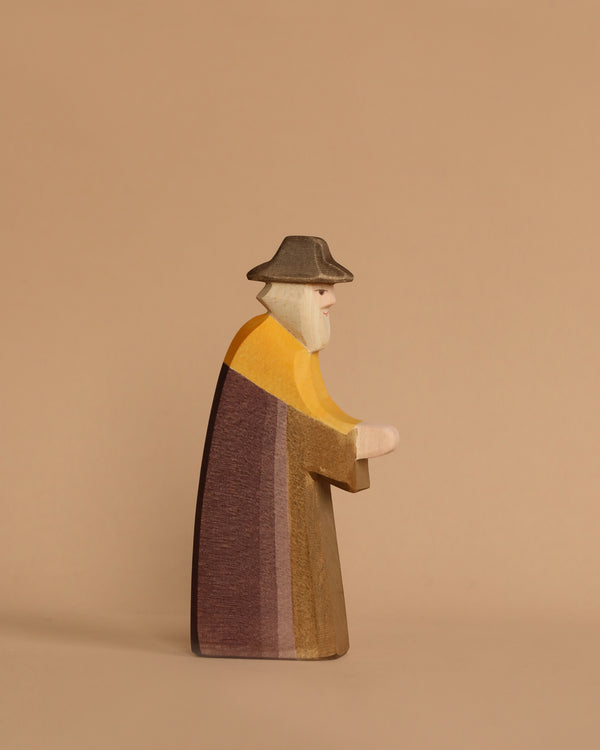 Against a plain beige background, the Ostheimer Joseph - Walking figure stands, depicting an elderly man with a beard wearing a hat and long multicolored coat. Handcrafted in the minimalist style typical of Ostheimer toys, it inspires imaginative play with its charming simplicity.