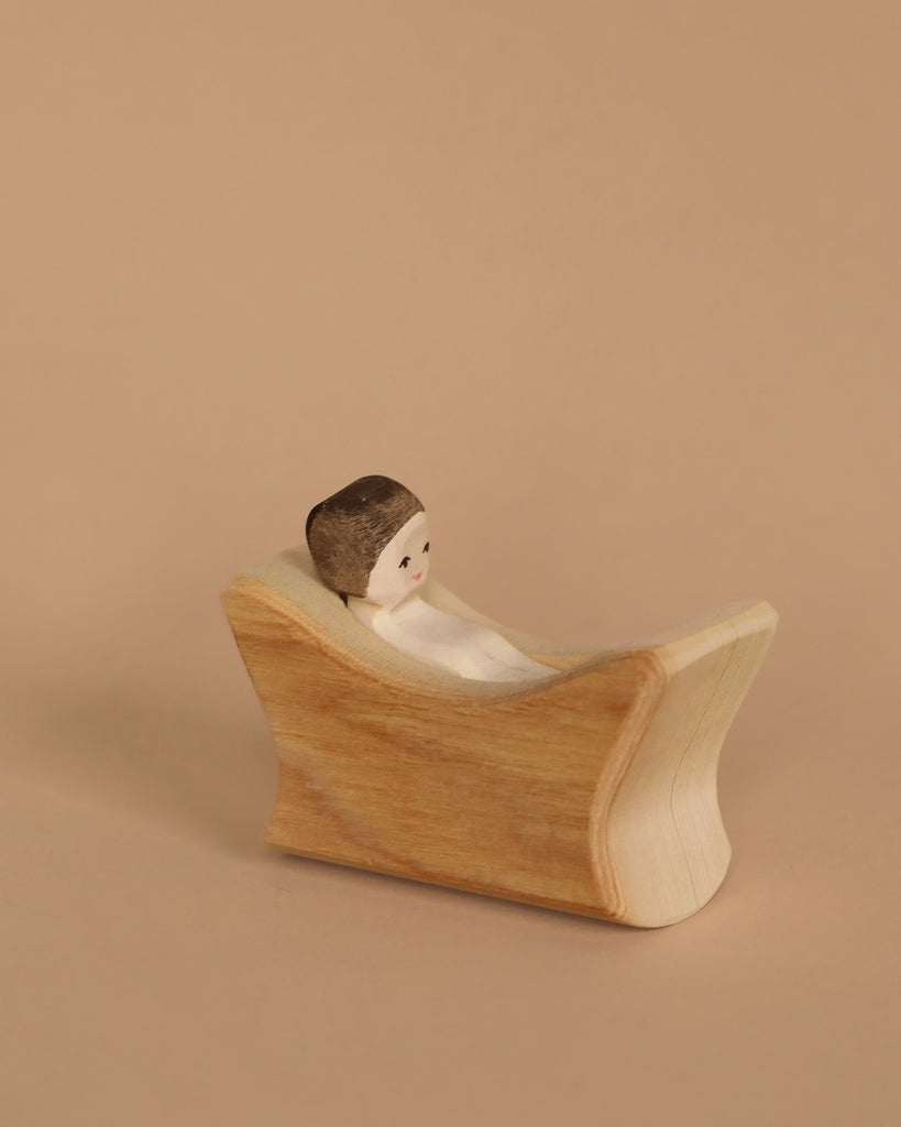 The Ostheimer Nativity Scene features a small wooden figure with a painted face, nestled inside a curved block like a cozy cradle, set against a neutral beige background, perfect for inviting imaginative play.