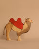 An Ostheimer Nativity Scene camel figurine, featuring a red painted saddle, stands against a beige background. Handcrafted from sustainable materials, this camel boasts a simplistic, stylized design that invites imaginative play.