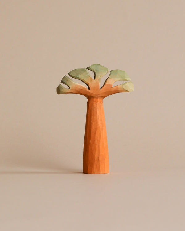 A Hand Carved Baobab Tree with a tall, tapering trunk and a flat, broad canopy featuring green leaves, set against an off-white background. Perfect for kids' room decor or as part of a wooden safari toys collection.